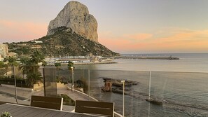 A coastal view with a prominent rock formation, a marina with boats, and a promenade with benches.