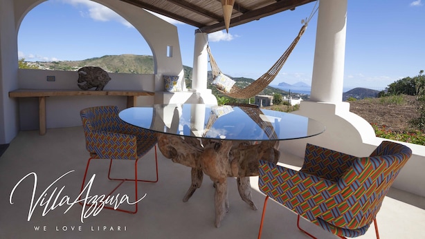 A round glass table with a wooden base, surrounded by patterned chairs, under a pergola with a view of hills. - Villa Azzura Lipari - Charm & Relax (Lipari)