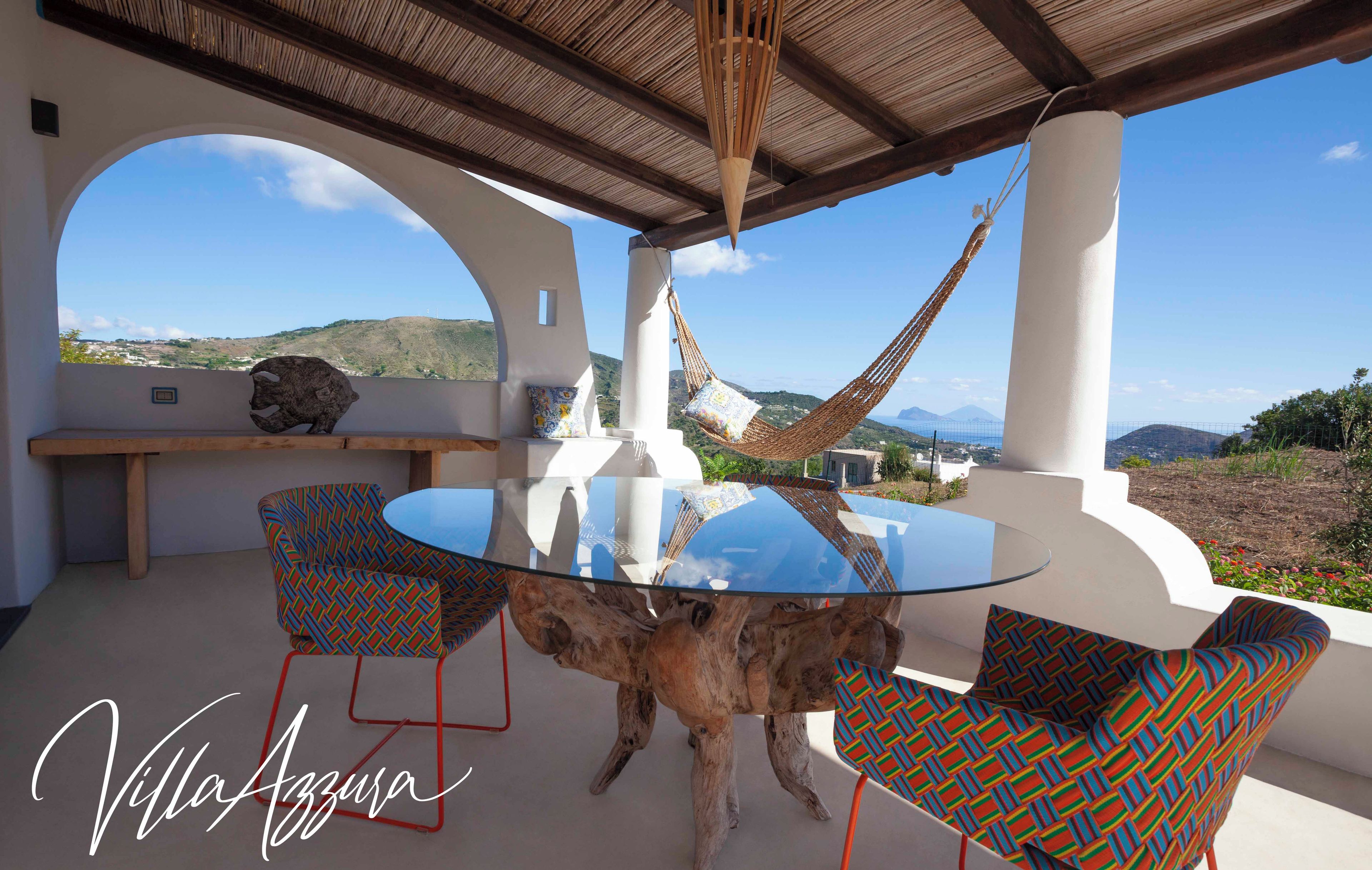 A round glass table with a wooden base, surrounded by patterned chairs, under a pergola with a view of hills.
