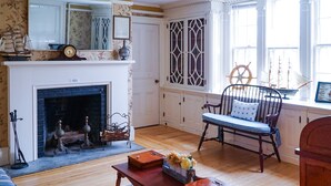 A room with a fireplace, a wooden floor, a chair with a cushion, a table with a vase, and a window with a ship wheel decoration. - Charming estate on Norwich Green, near casinos (Norwich)