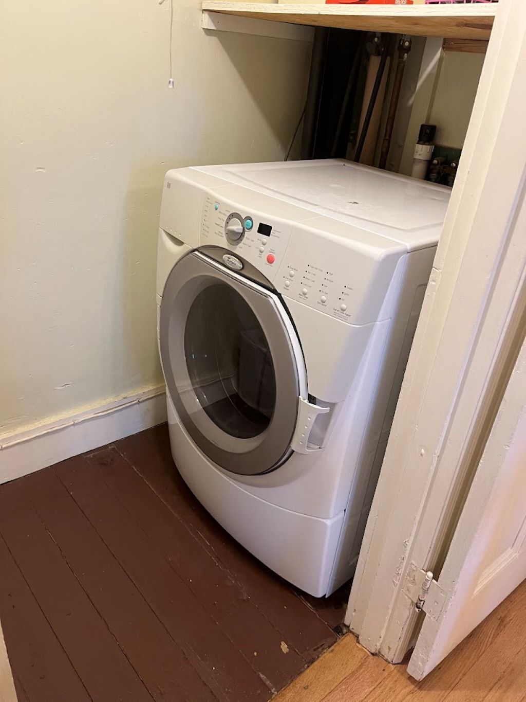 Lavadora blanca de carga frontal, compacta, en un pequeño cuarto de lavado con piso de madera y una puerta blanca.