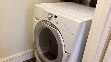 Lavadora blanca de carga frontal, compacta, en un pequeño cuarto de lavado con piso de madera y una puerta blanca.