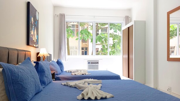 A hotel room with two beds, a large window with a view of greenery, a ceiling fan, and a wall-mounted air conditioner. - TC308 Aconchegante Studio na Praia (Rio de Janeiro)