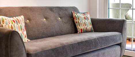 A grey sofa with two patterned cushions and a window with a view of a patterned chair outside.