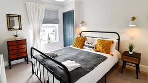 A bedroom with a bed, a dresser, a nightstand, a window with curtains, and a wall-mounted light fixture. - Just Stay Wales - Hadland House Mumbles (Swansea)