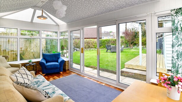A sunlit room with a large sliding glass door leading to a garden, a blue armchair, and a wooden table with a vase of flowers. - Just Stay Wales - Hadland House Mumbles (Swansea)