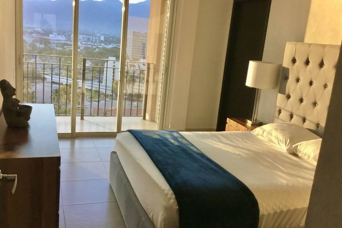 Une chambre d’hôtel avec un lit, une table de chevet, une lampe, une statue et une vue sur le paysage urbain, visible à travers de grandes fenêtres.