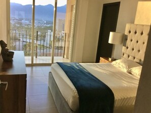 A hotel room with a bed, a nightstand, a lamp, a statue, and a view of a cityscape through large windows. - Grand Venetian Vallarta (Puerto Vallarta)