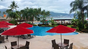 A pool area with red umbrellas and lounge chairs, surrounded by palm trees and a view of the city and mountains.