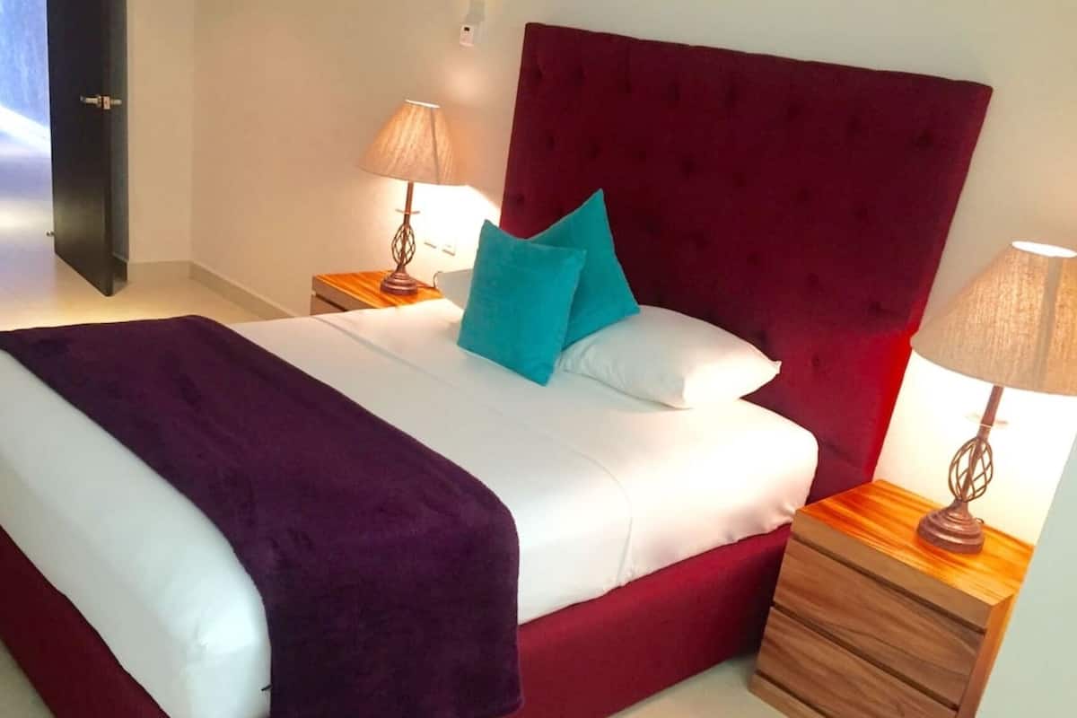 Un lit avec une tête de lit rouge, une literie blanche et deux coussins turquoise. On y trouve deux tables de chevet en bois avec des lampes.