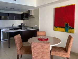 Cozinha moderna com armários escuros, pia de aço inoxidável e micro-ondas. Há uma área de jantar com mesa redonda e cadeiras listradas. Um quadro abstrato e colorido está na parede.