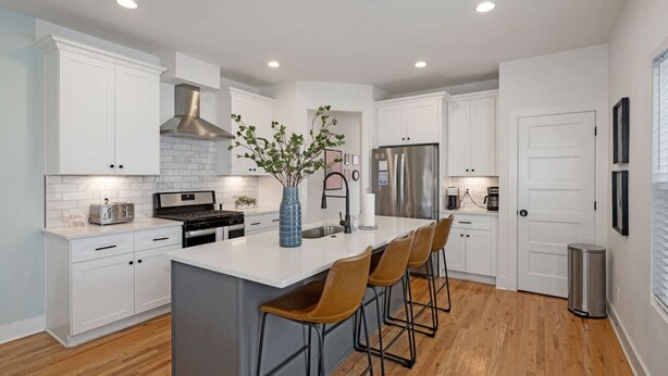 A modern kitchen with white cabinetry, a central island with bar stools, stainless steel appliances, and a decorative vase. - Modern Townhouse: 3-Bed Haven, Vibrant Pink Door! (Nashville)