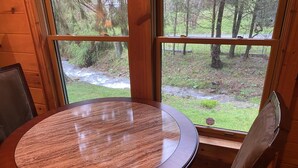 Una mesa de madera redonda con vistas a un bosque y un arroyo a través de amplios ventanales.