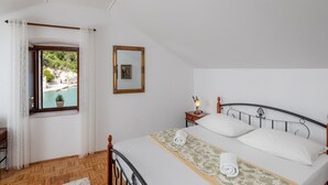 Une chambre à coucher comprenant un lit, une table de chevet, une lampe, une fenêtre donnant sur un plan d’eau et un tableau encadré accroché au mur.