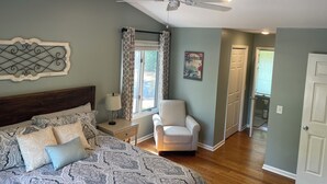 Ein Schlafzimmer mit Bett, Sessel, Deckenventilator, Fenster mit Vorhängen und einer Tür.