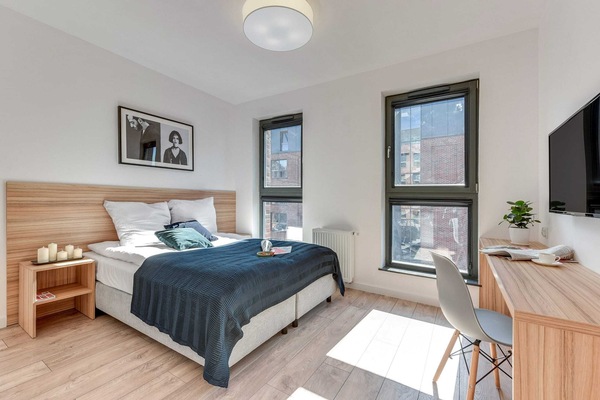 A bright bedroom featuring a comfortable bed with blue bedding, a small desk, and large windows providing natural light. The space is modern and functional, ideal for work or relaxation.