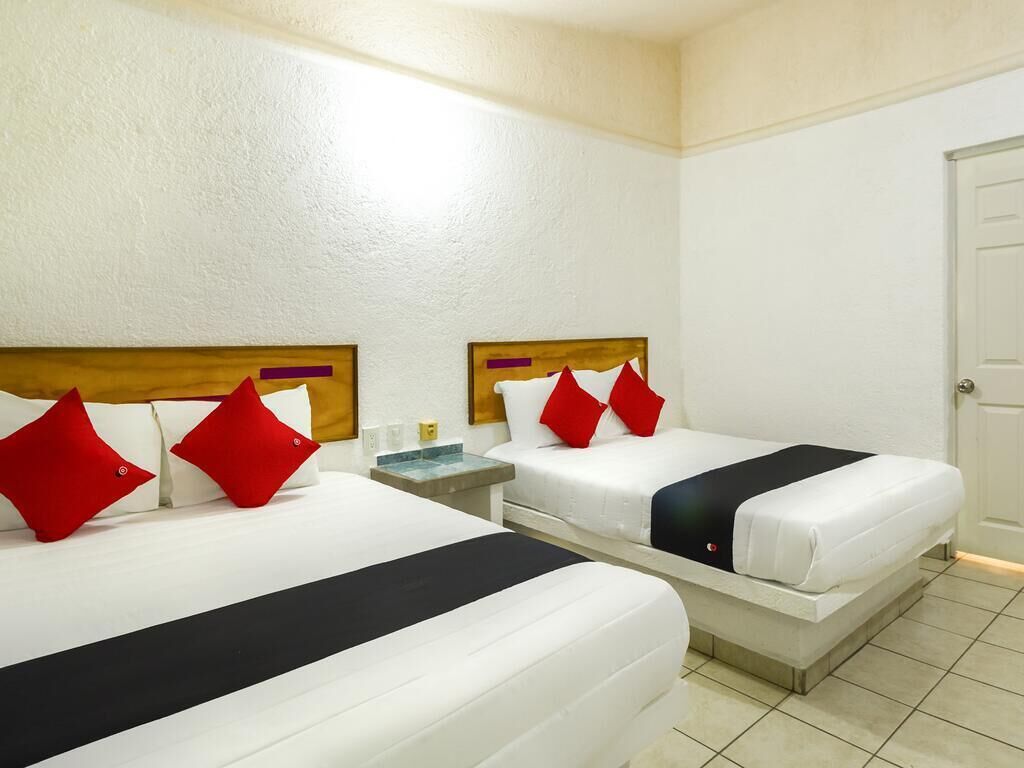 A hotel room with two beds, each with white bedding and red pillows, a wooden headboard, and a bedside table with a lamp.