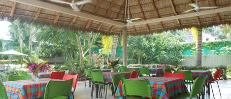 An outdoor dining area with thatched roof, green chairs, and tables covered with colorful tablecloths.
