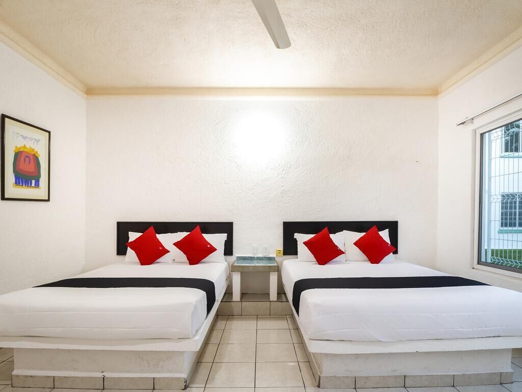 A hotel room with two beds, each with white bedding and red pillows, a small table between the beds, and a window on the right side.
