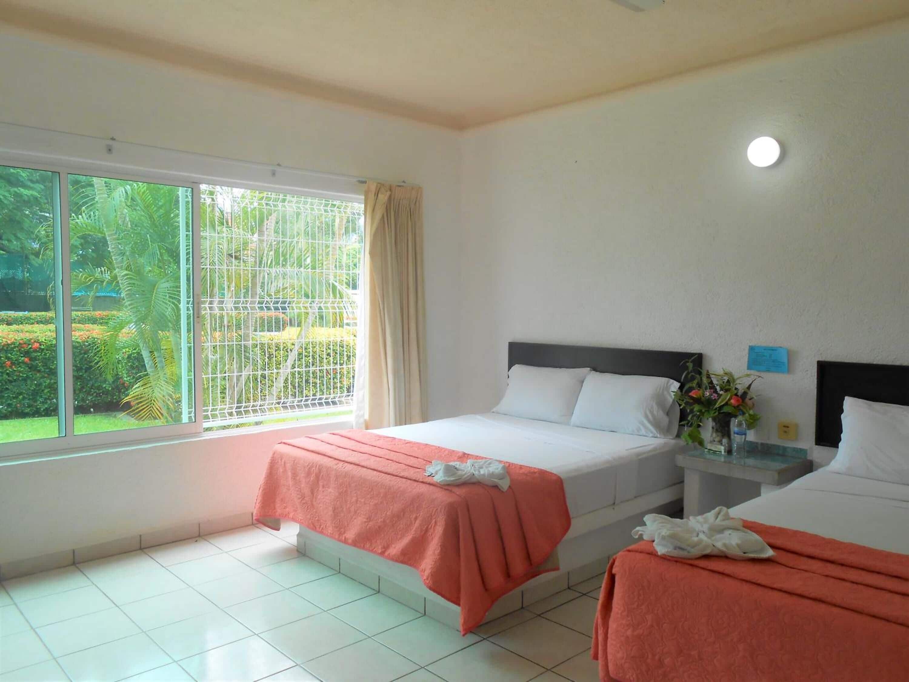 A hotel room with two beds, a window with a view of greenery, and a small table with a vase of flowers.