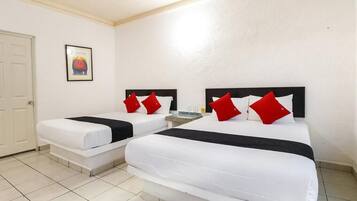 Habitación de hotel con dos camas, cada una con ropa de cama blanca y almohadas rojas. Se observa una puerta y un cuadro en la pared.
