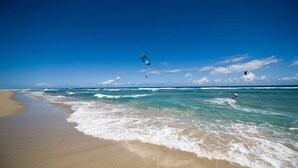 En strand med klart himmel, bølger og en person, der laver kitesurfing.