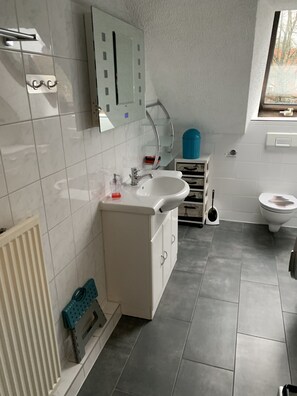 Un bagno moderno con lavandino, specchio e water bianchi. Una finestra offre luce naturale e un termosifone si trova sul lato sinistro.