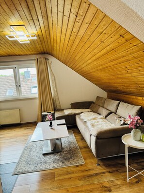 Ein gemütliches Schlafzimmer im Loft-Stil mit schrägem Dach, einem großen Fenster, einer Couch, einem Couchtisch und einem kleinen Beistelltisch mit Blumen.