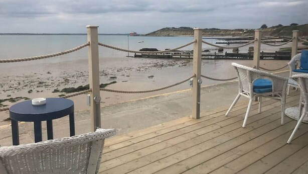 A wooden deck with wicker chairs and a small table, overlooking a beach with a rope barrier and distant boats. - Cosy traditional cottage with parking and garden (Ryde)