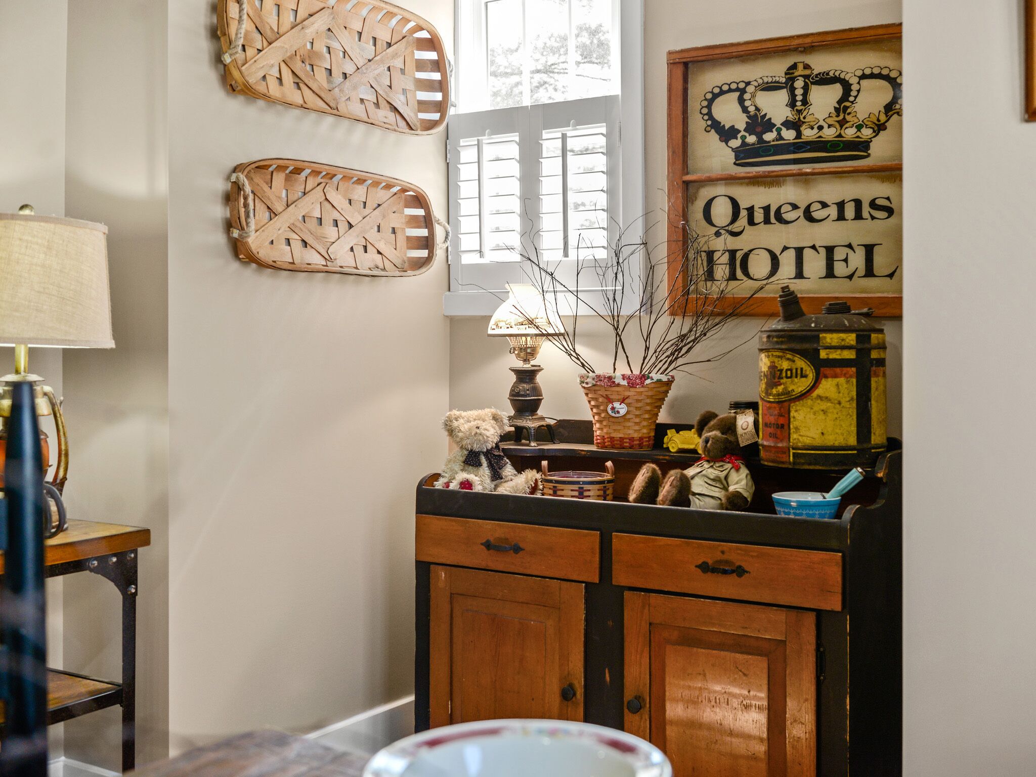 Une chambre au style rétro, avec un meuble en bois, une lampe, un panier en osier, un ours en peluche et une enseigne indiquant « Queens Hotel ».