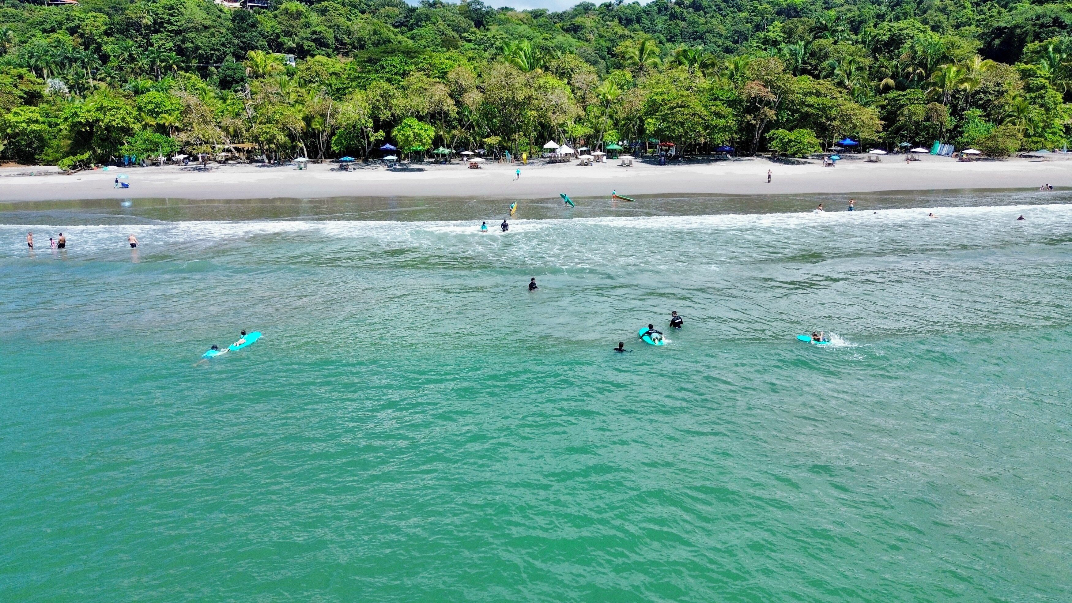A beach with clear turquoise water, people surfing, and a sandy shore with beach umbrellas.