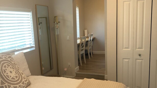 Ein Schlafzimmer mit einem großen Bett, einem Nachttisch, einem Spiegel, einem Fenster mit Jalousien und einer Tür zu einem Flur mit Schreibtisch und Stühlen.