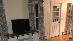 Ein Wohnzimmer mit Fernseher auf einem weißen Schrank, gemusterten Vorhängen, Holzboden und grauem Teppich.
