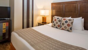 A hotel room with a large bed, a wooden headboard, a bedside lamp, a TV on a wooden cabinet, and a patterned throw blanket. - CASA DEL MAR BEACH RESORT (Ormond Beach)