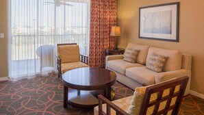 Habitación de hotel con sofá, dos sillones, una mesita, lámpara y un ventanal grande con cortinas.
