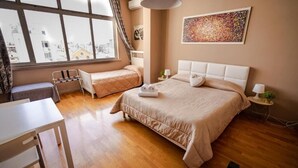 A bedroom with two beds, a desk, a chair, a radiator, and a window with a view of buildings. - Dolci Emozioni Room 82 (Bari)