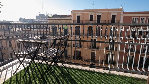 Un balcón con mesa y sillas, rodeado de baranda metálica, con vista a un paisaje urbano de edificios.