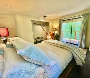 A hotel room with two beds, a window with a view of greenery, a small table with a lamp, and a chair.