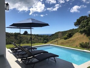 A swimming pool with lounge chairs and an umbrella, surrounded by greenery and hills. - The Ranch At La Joya is a three bedroom modern farm house at Tres Palmas Estates (Rincón)