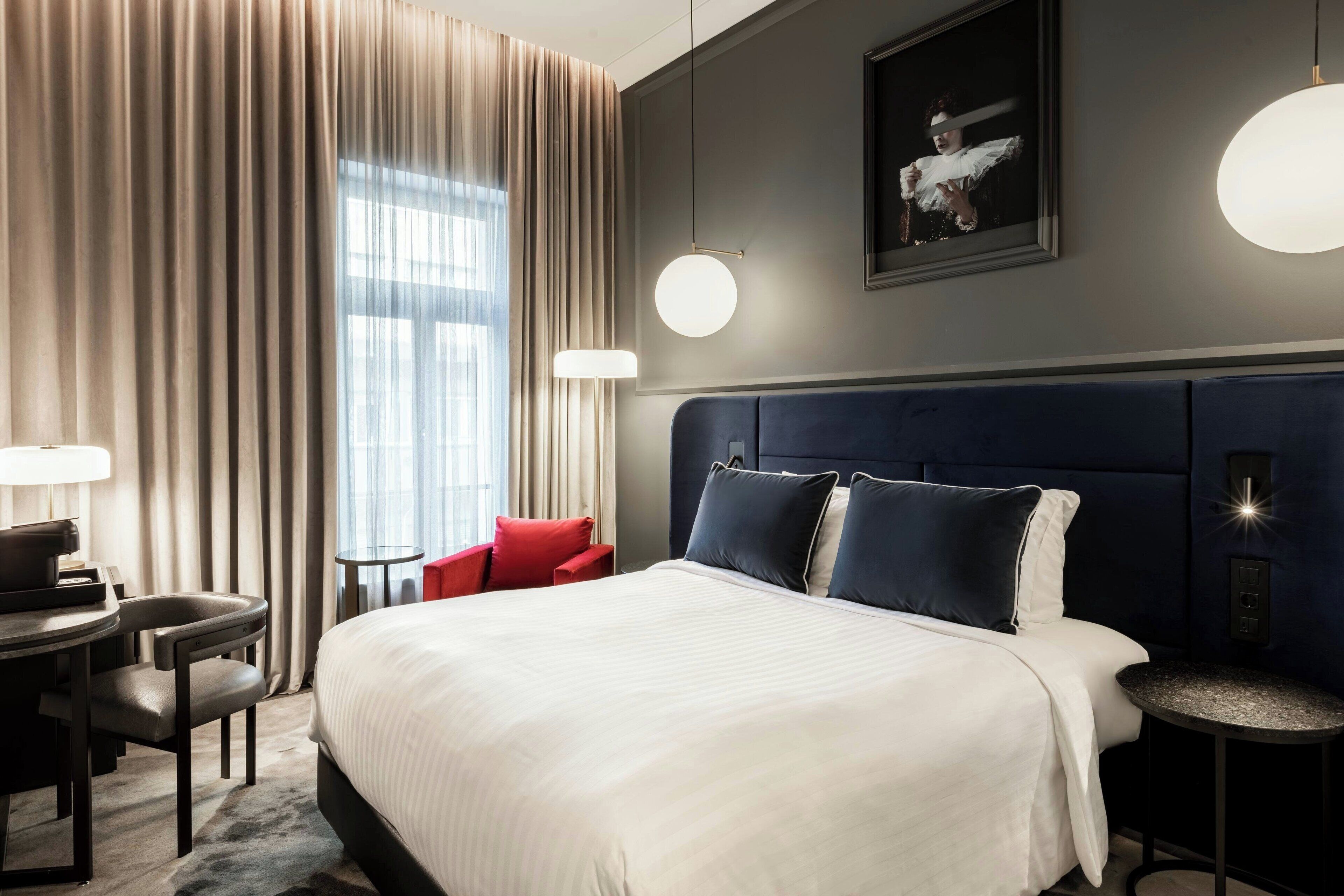 A hotel room with a large bed, a desk with a chair, a small table, and a red chair. There is a window with curtains, a wall-mounted light, and a framed picture.