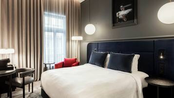 A hotel room with a large bed, a desk with a chair, a small table, and a red chair. There is a window with curtains, a wall-mounted light, and a framed picture.