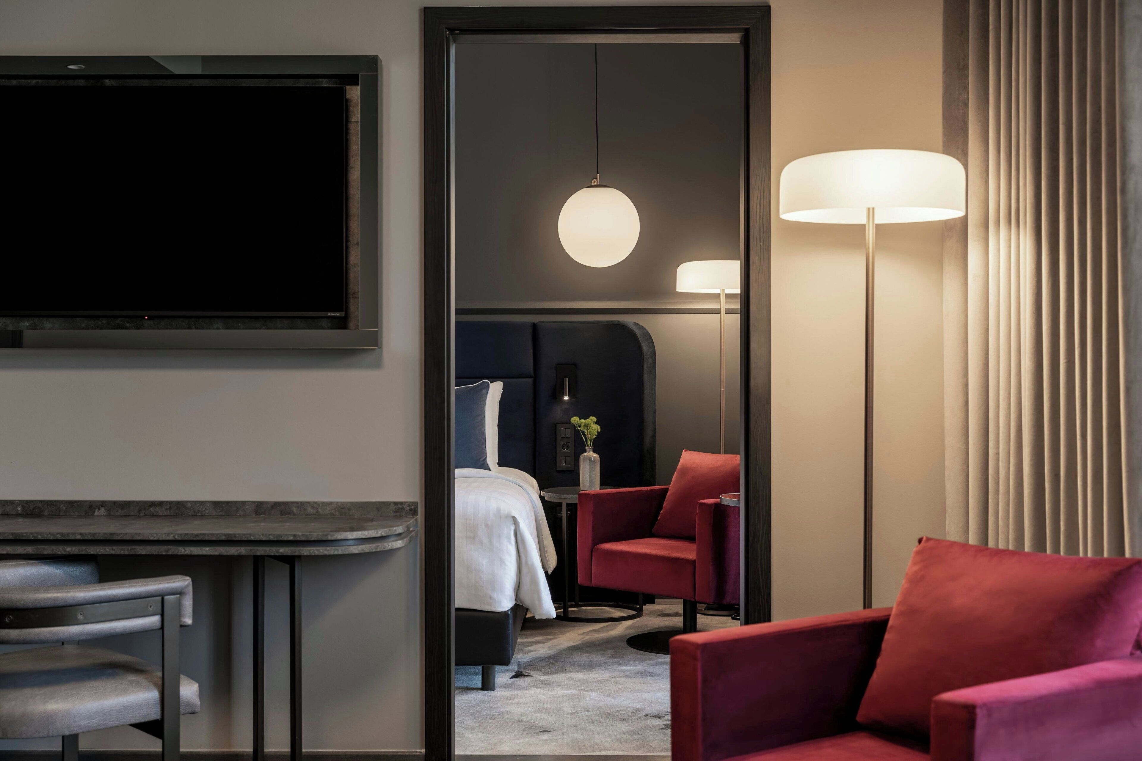 Una habitación de hotel con una cama, un sillón rojo, una mesita, un televisor y una lámpara de pie.