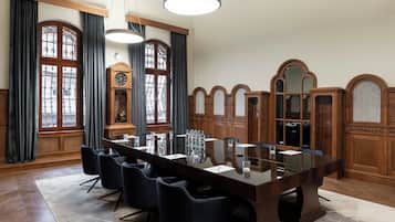 Un salón de conferencias con una larga mesa, sillas negras, paneles de madera y grandes ventanales con cortinas.