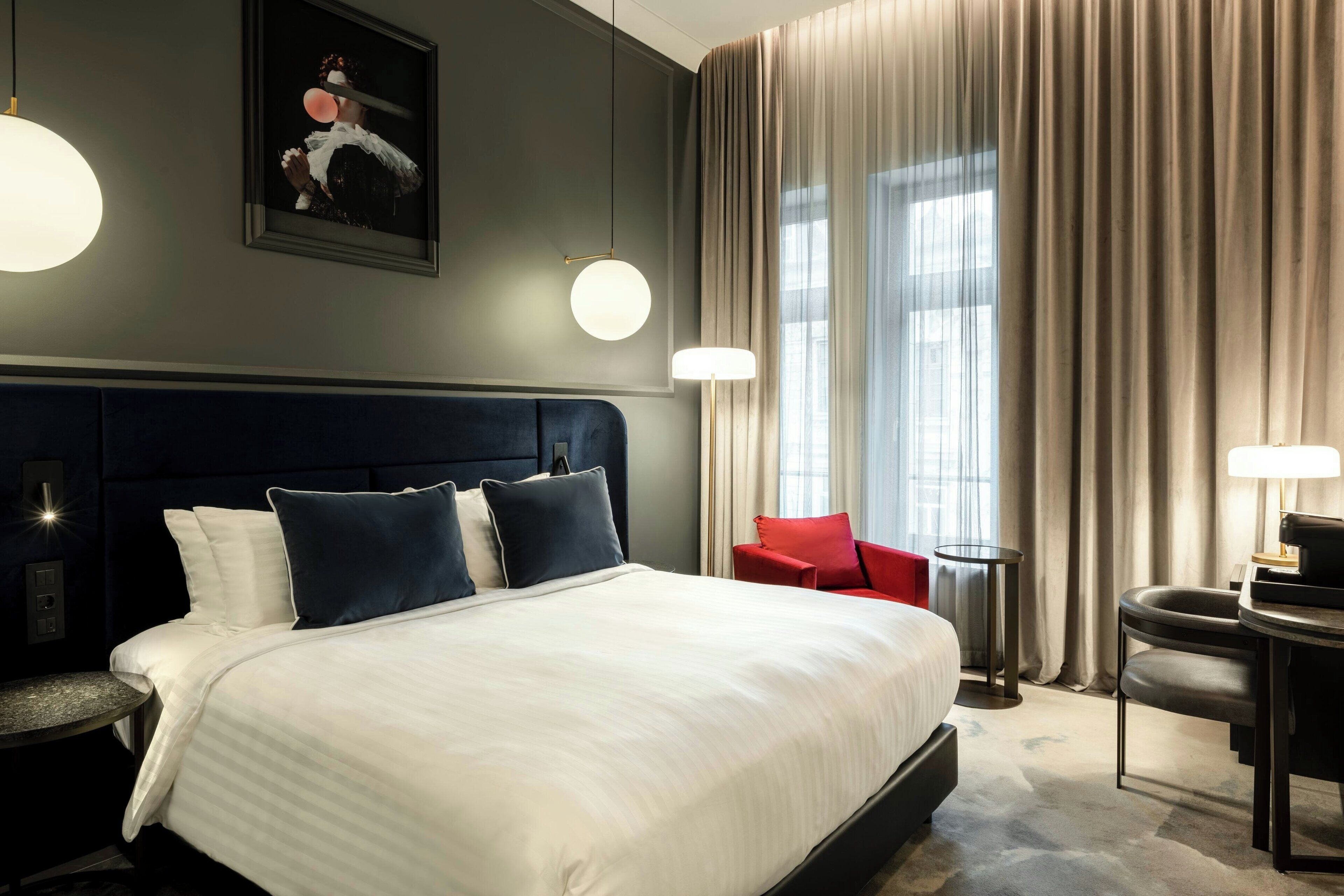 Una habitación de hotel con una cama grande, una silla roja, una mesita y un cuadro en la pared.