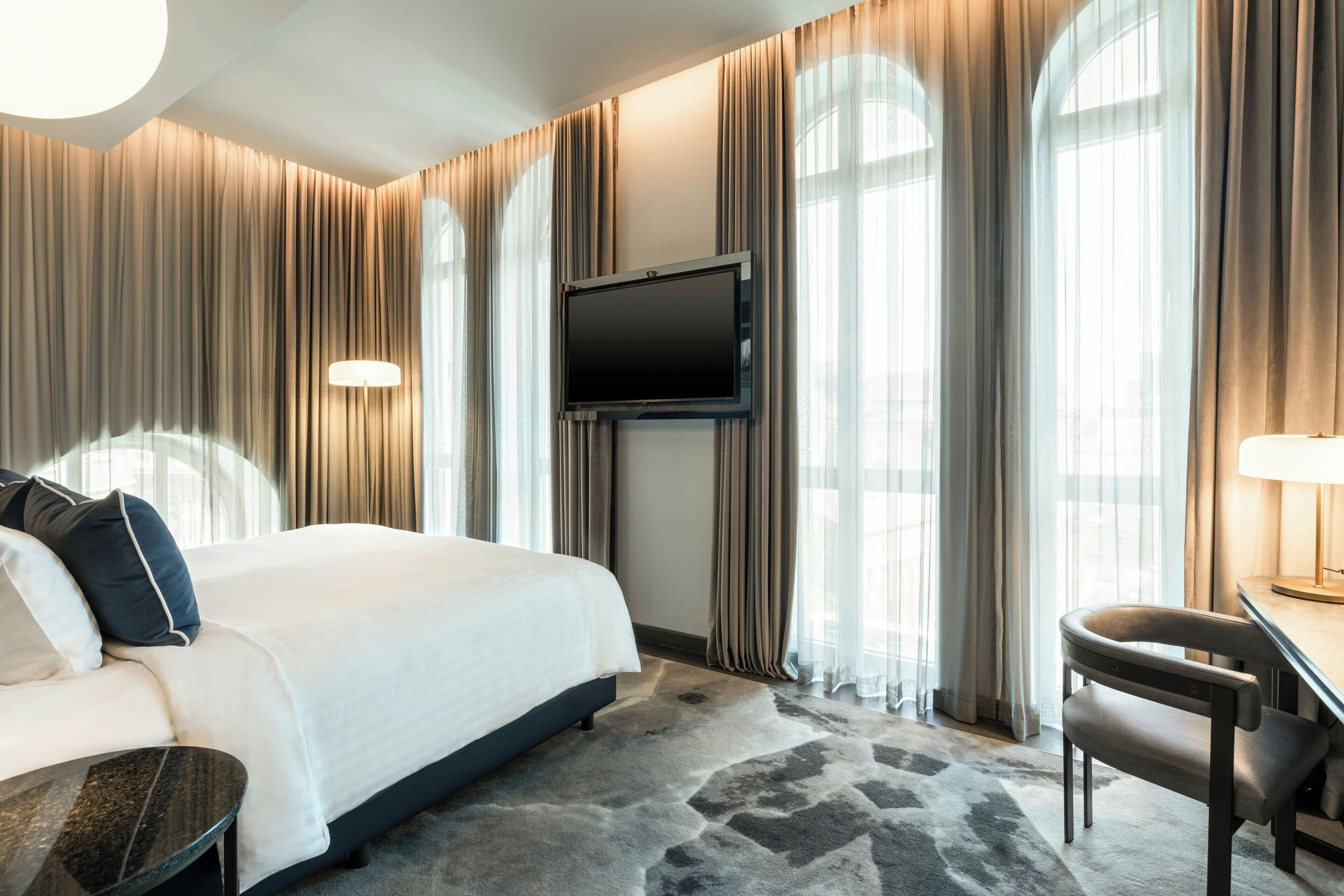 A hotel room with a large bed, a flat-screen TV mounted on the wall, a desk with a chair, and a large window with curtains.
