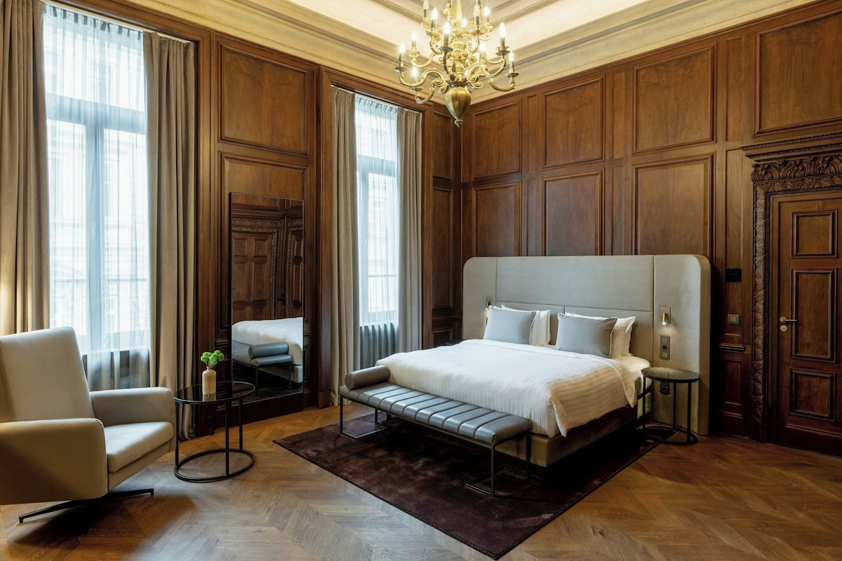 A spacious bedroom with a large bed, a sitting area with a chair and a small table, and a large window with curtains.