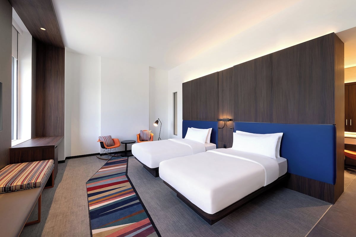 A modern hotel room with two beds, a bench, a colorful striped rug, and a large window.
