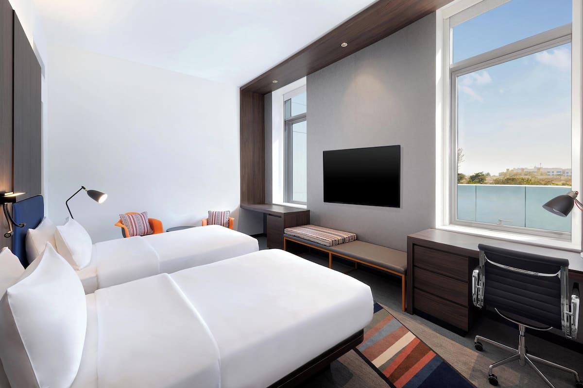 A modern hotel room with two beds, a desk, a TV, and a large window offering a city view.