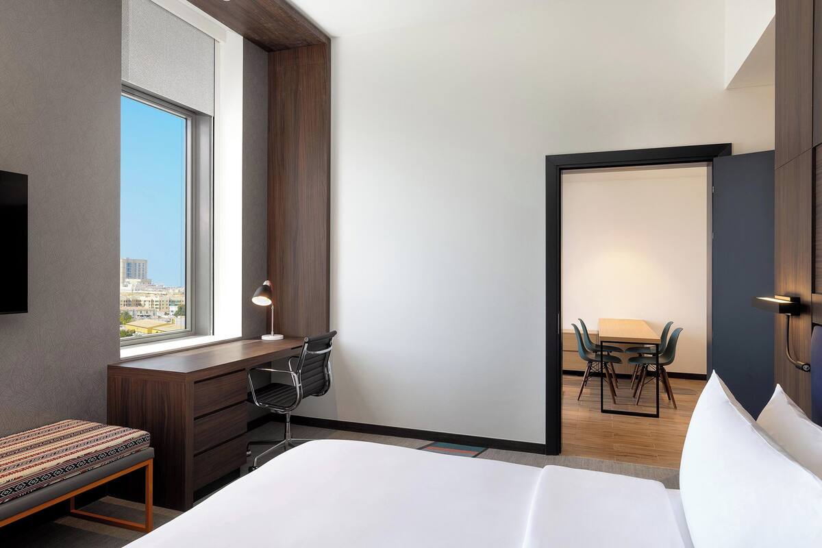 A modern hotel room with a large bed, a desk with a chair, a small bench, a dining area with a table and chairs, and a television mounted on the wall.