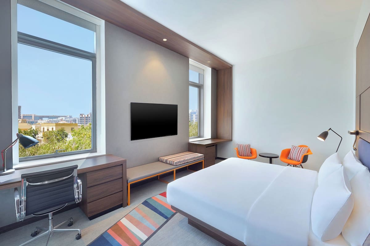 A modern hotel room with a large bed, a desk, a TV mounted on the wall, and a view of the city through large windows.